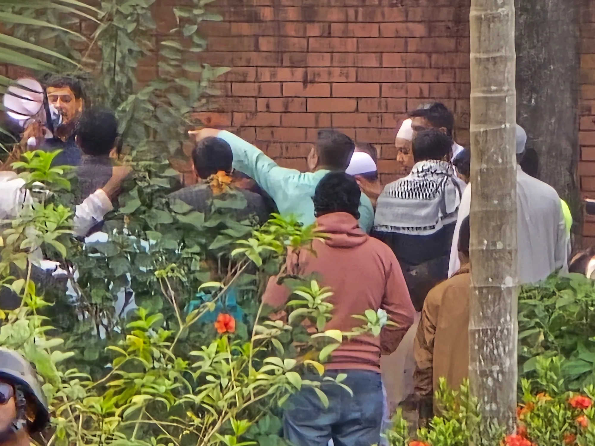 Men crowd together beside a red brick wall behind flowering shrubs; one man in the background speaks into a megaphone.