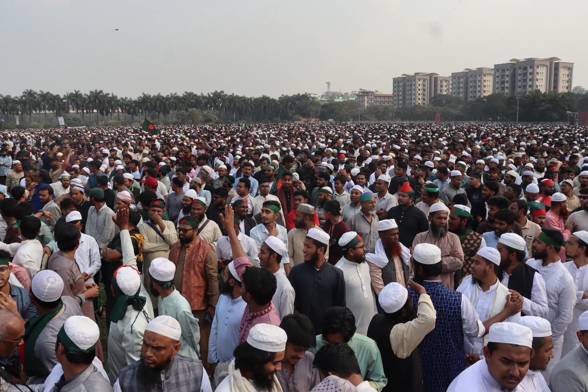A dense crowd of men, most in white prayer caps, stands on open ground; some raise their hands; apartment blocks stand on the horizon.