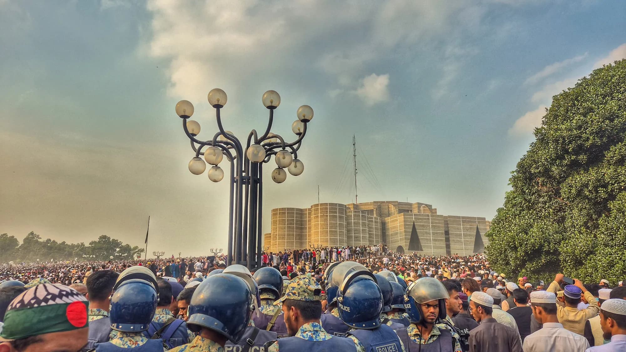 Rows of helmeted security personnel in the foreground face a large crowd; the parliament building and a tall lamp post stand behind.