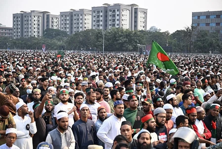 A dense crowd of men, many in white prayer caps, stands in rows on open ground before apartment blocks; one national flag is raised.