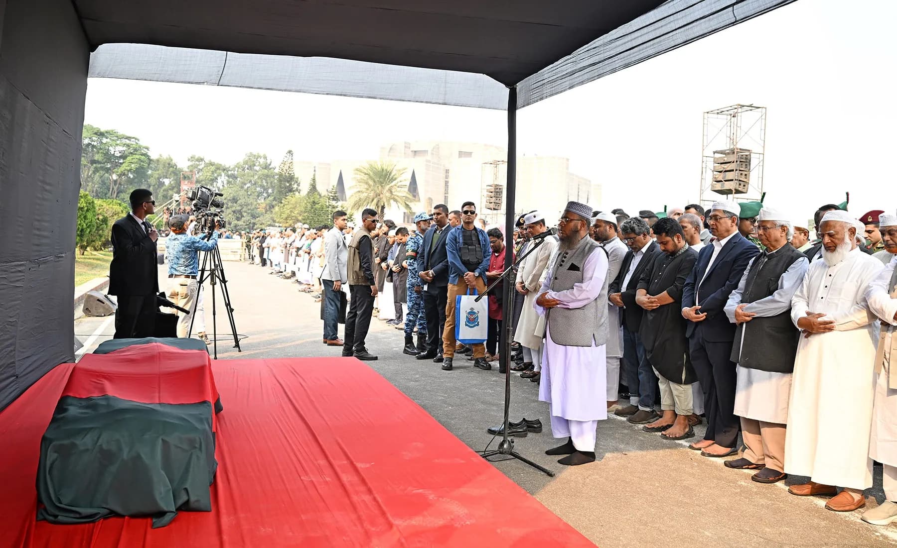 Men stand in a line in prayer under a canopy; before them a coffin covered with the national flag rests on a red carpet.