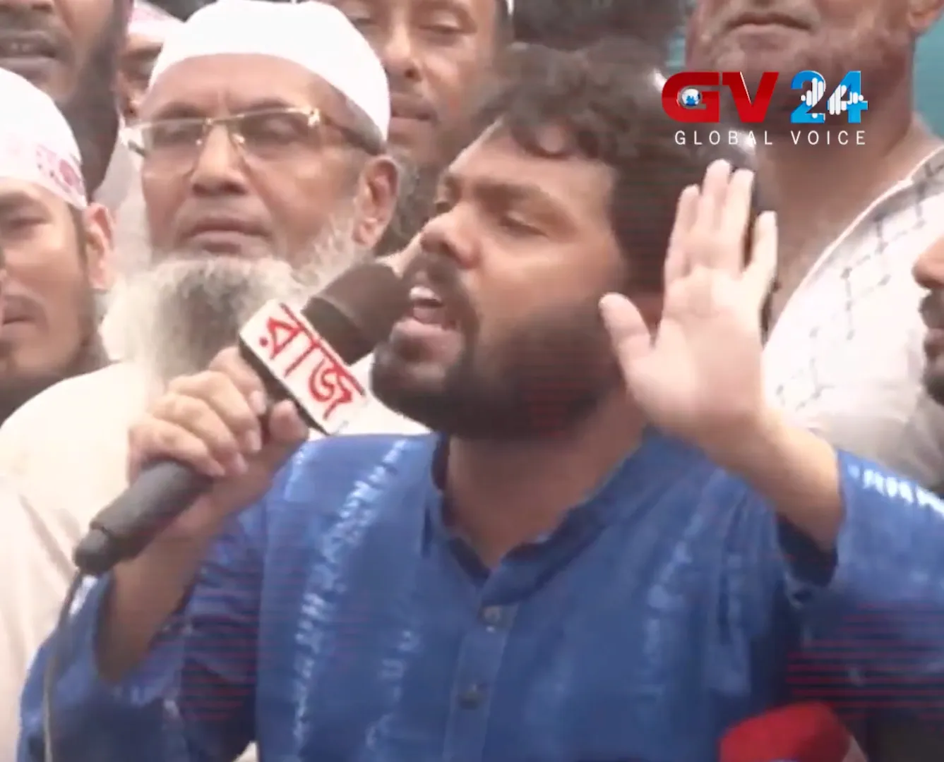 A bearded man in a blue panjabi speaks into a microphone and raises his open hand, with an older bearded man in a white cap behind him.