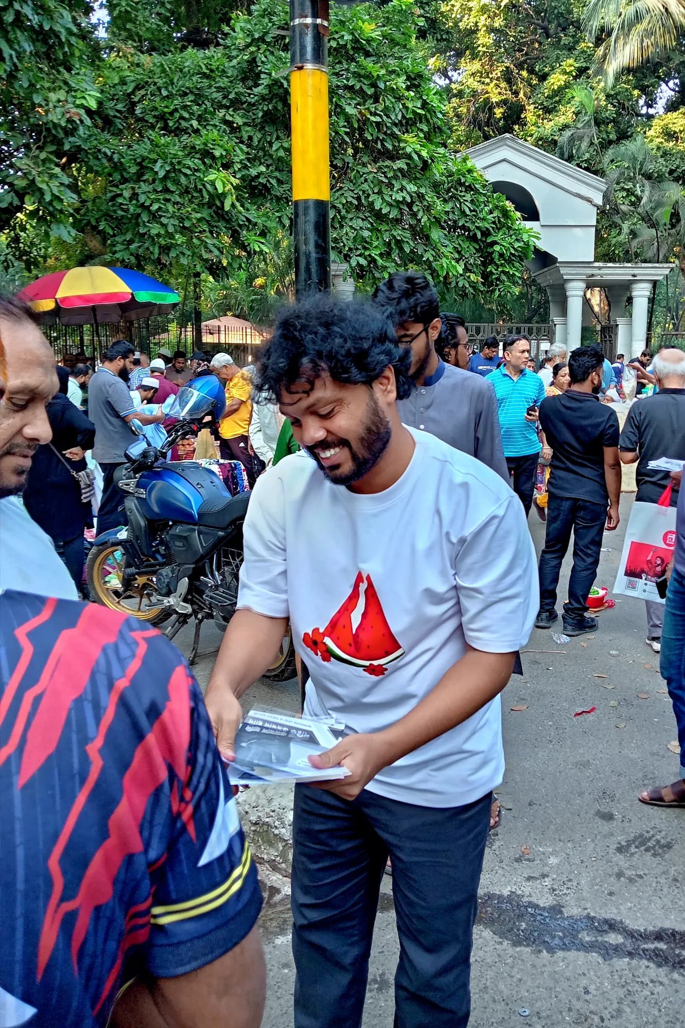 A smiling man with curly hair, in a white T-shirt printed with a red shape, holds out leaflets to a man in a striped shirt on a busy pavement.