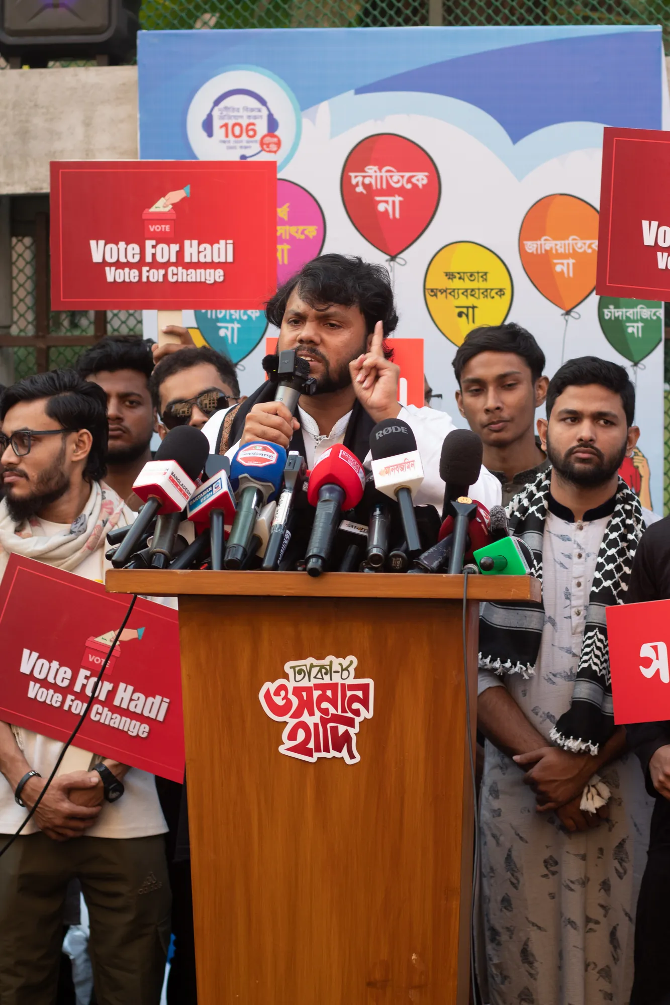 A bearded man with curly hair raises a finger as he speaks into a cluster of microphones at a lectern, with young men holding red placards around him.