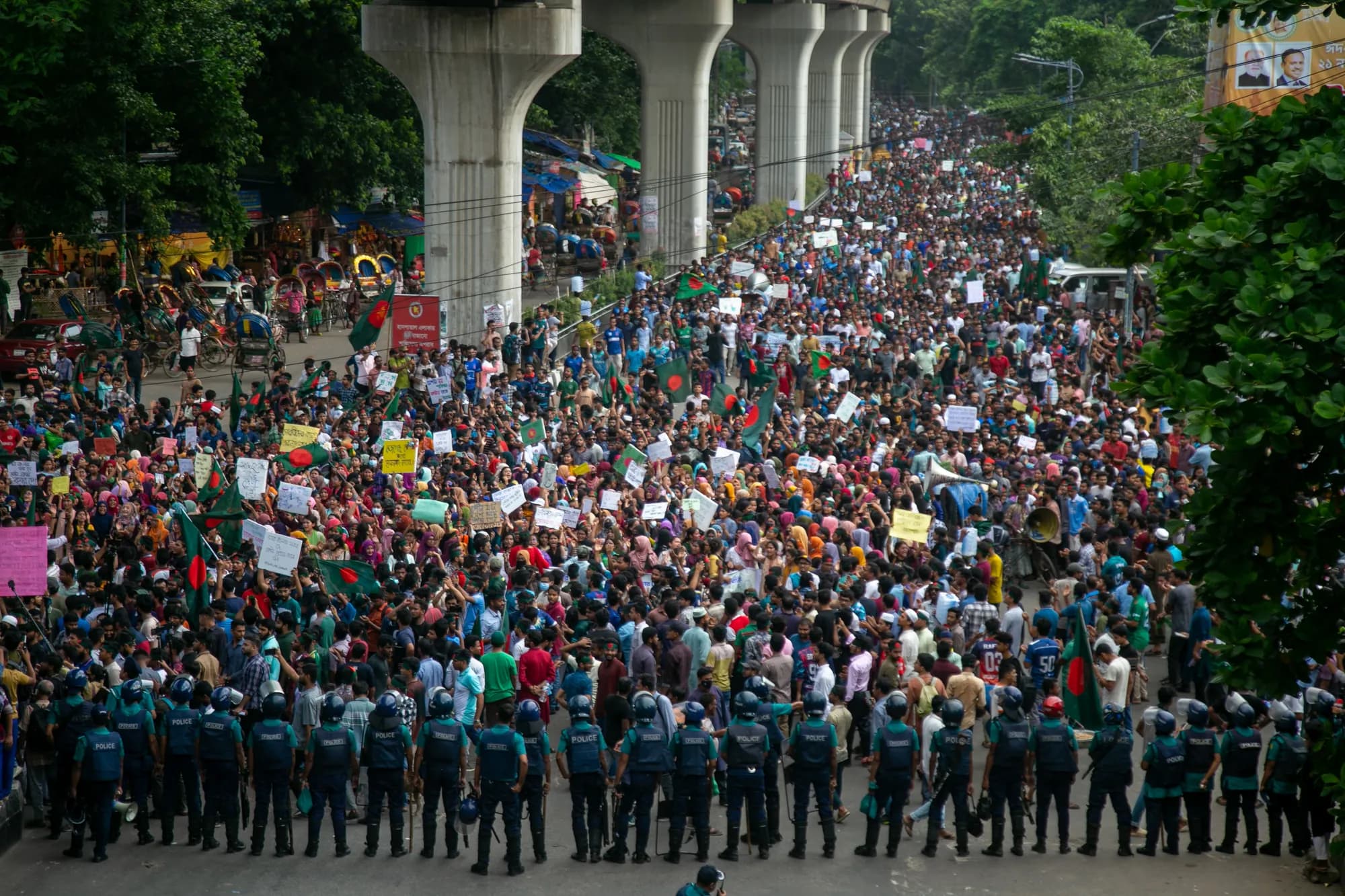 A huge crowd with placards and flags fills a road under the pillars of an elevated railway; a line of police in blue stands across the front.