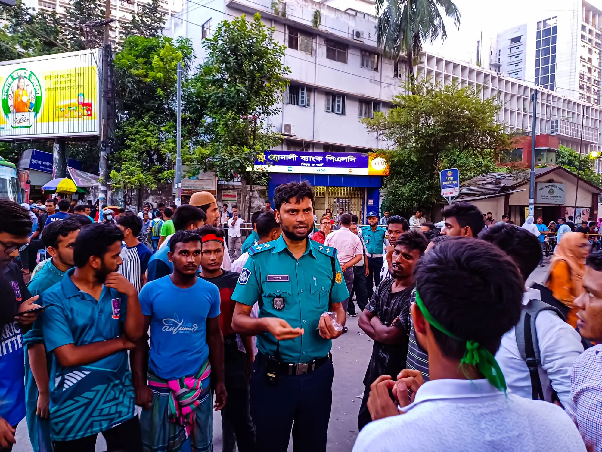 A police officer in a blue uniform gestures as he talks with a group of young men, one wearing a green headband, on a city street.