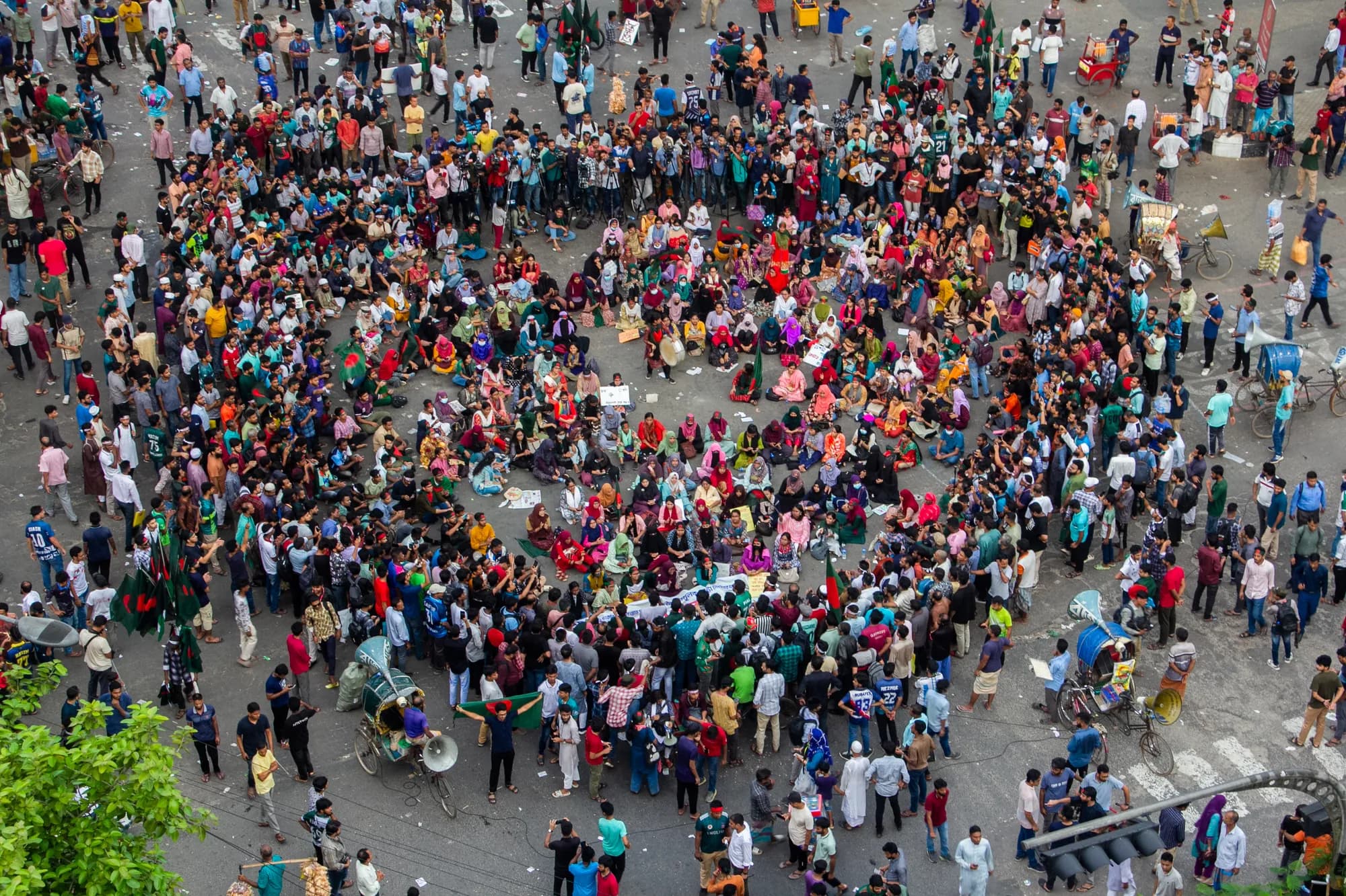 Seen from above, hundreds of people fill a road junction, some seated in a ring, with rickshaws at the edges.