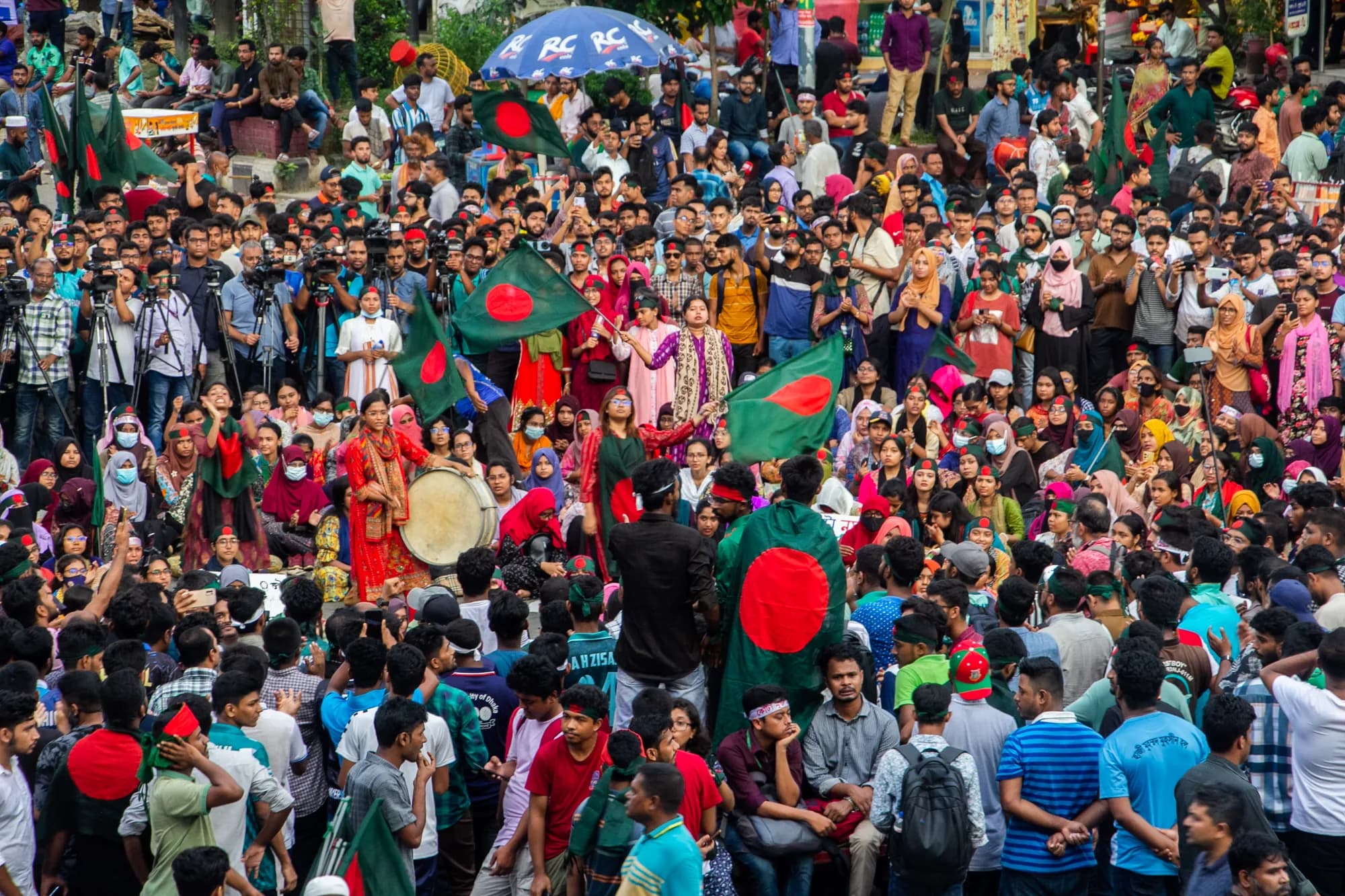 A dense crowd of young men and women, many with Bangladeshi flags, sits and stands around a drummer in the middle of a road.