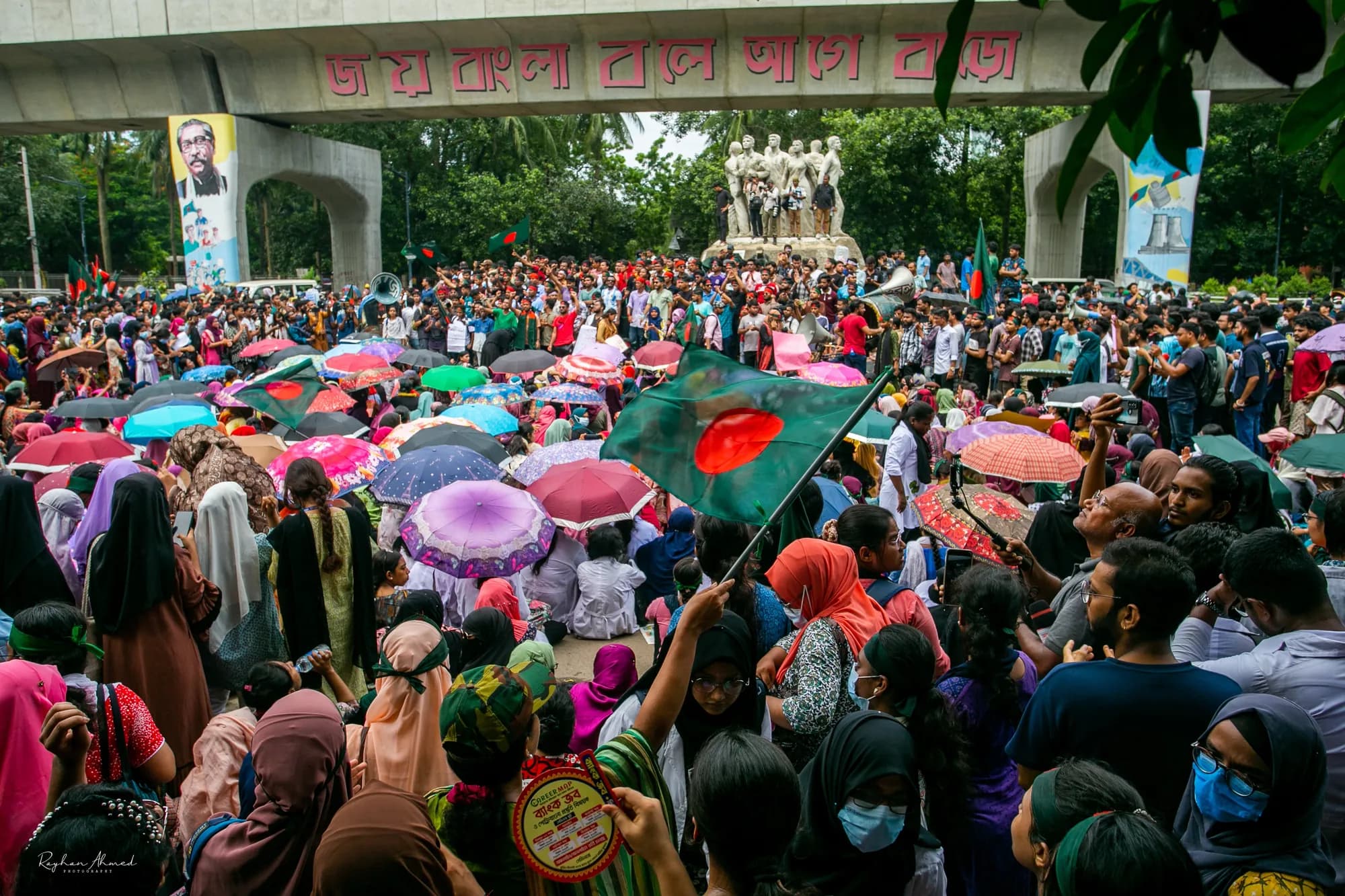 A crowd with coloured umbrellas and a Bangladeshi flag gathers below a bronze sculpture of figures under an arched gateway.
