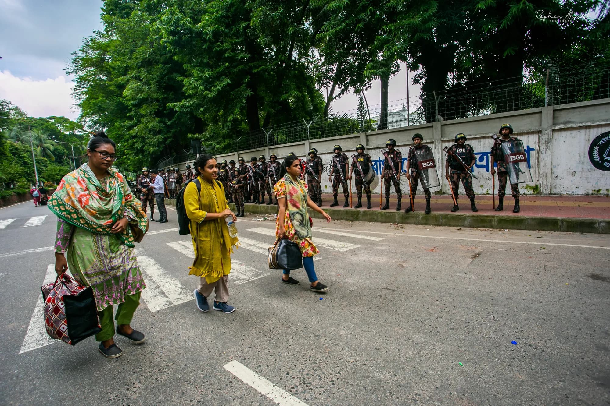 Three women in colourful clothes carry bags across a pedestrian crossing; a line of helmeted police stands along a wall behind them.