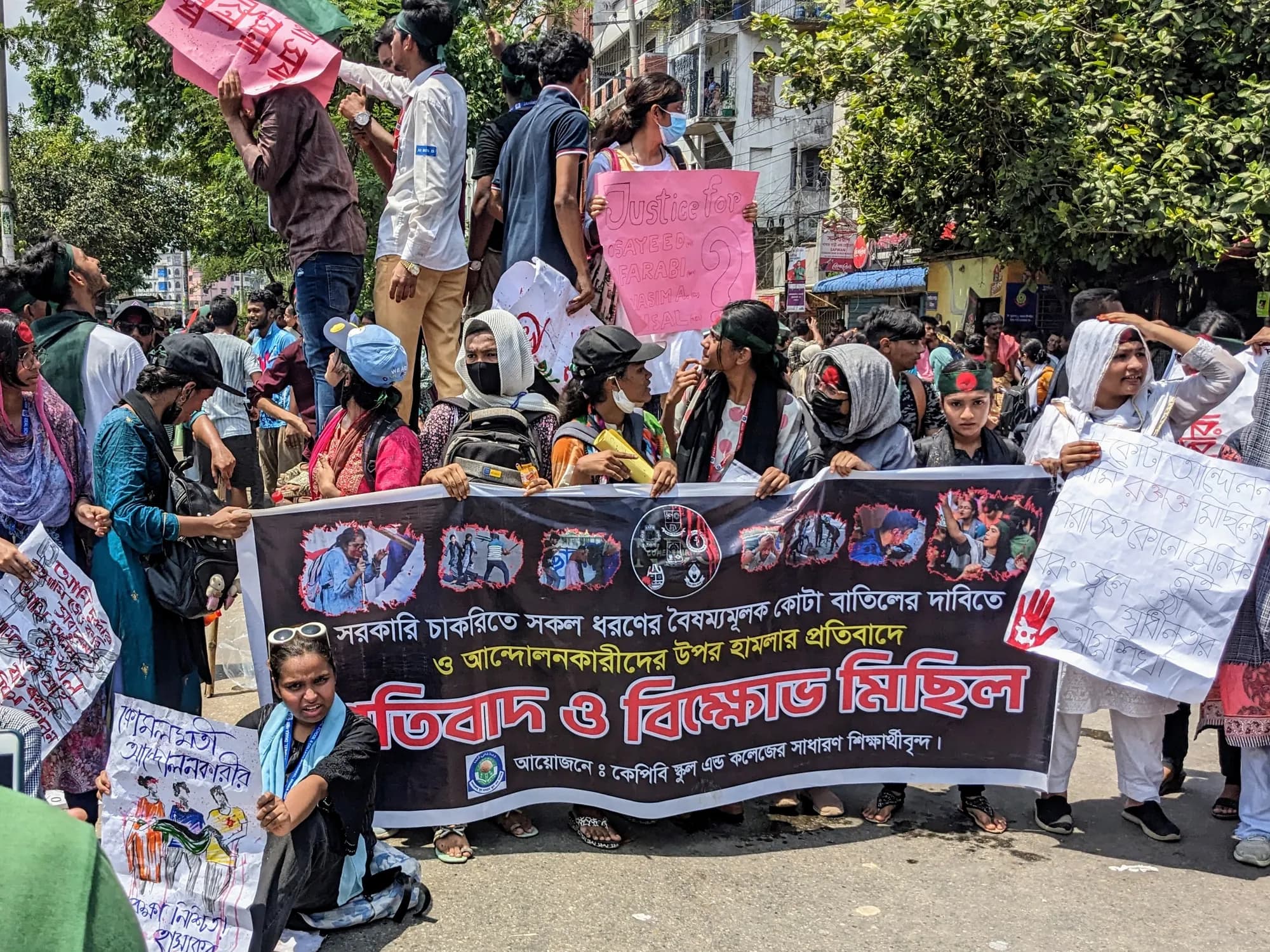 Young people stand behind a dark banner with red and white Bangla text and printed photographs, holding placards on a city street.