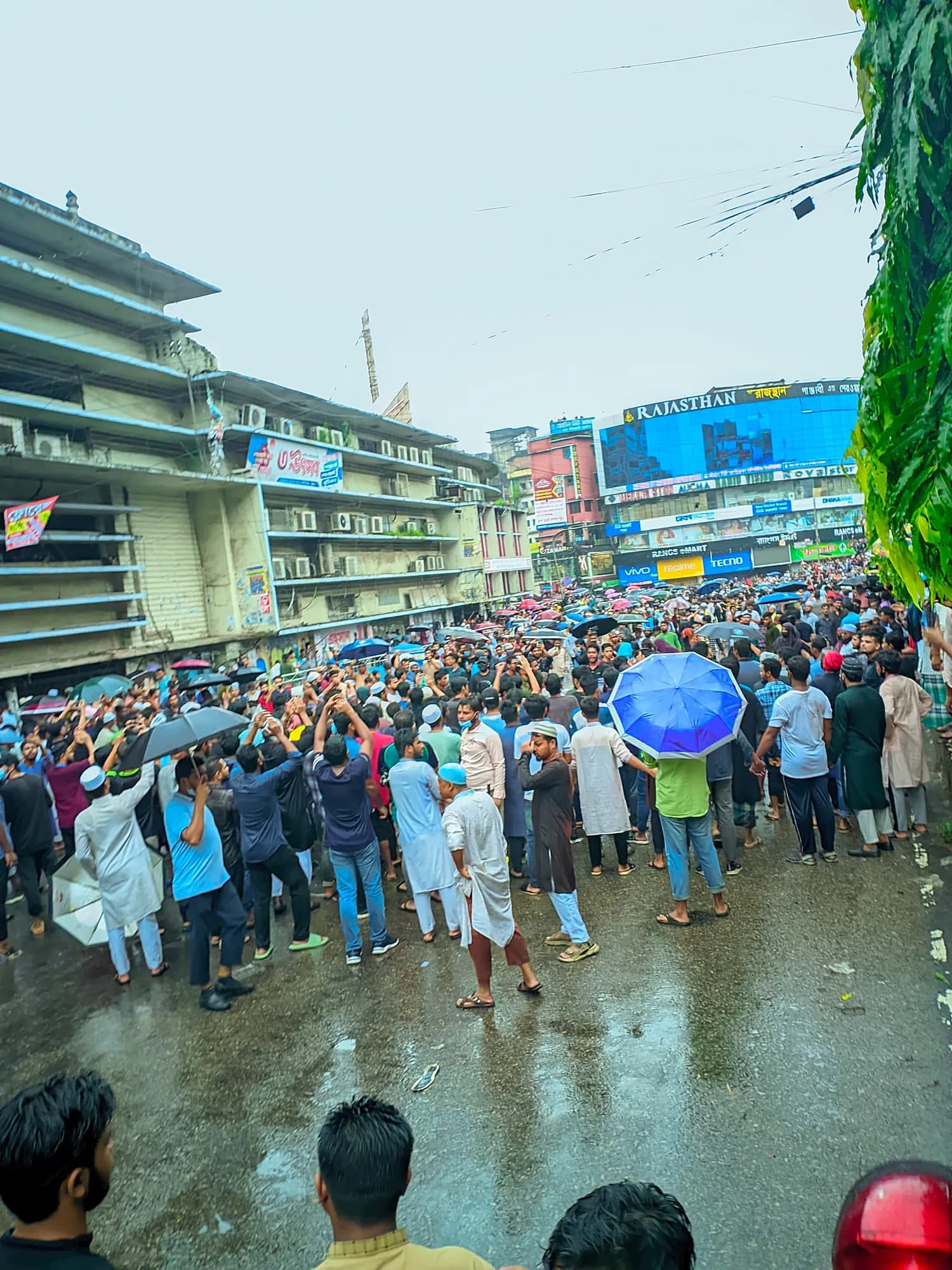 A crowd, some with umbrellas, gathers on a wet road between shop buildings with large billboards.