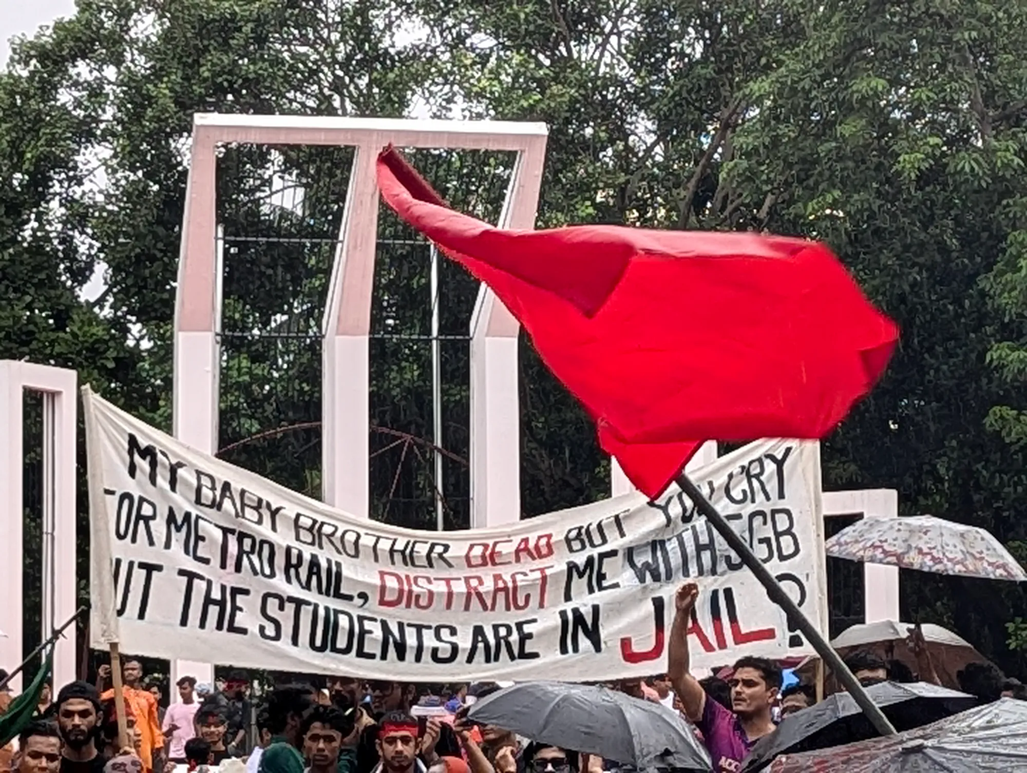 A white cloth banner with hand-painted black and red English lettering and a red flag are held up before the white pillars of a monument.