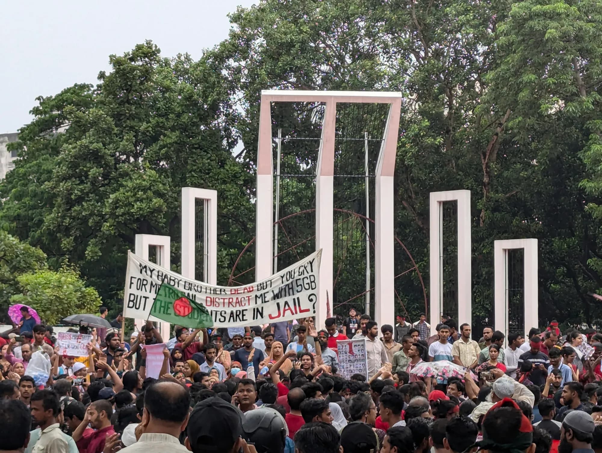 A crowd with umbrellas, flags and banners gathers before the tall white frames of a monument, with trees behind.