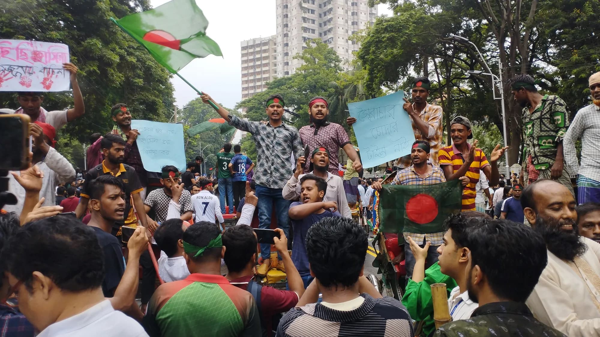 Young men wave a Bangladeshi flag and hold pale blue placards in a crowd under trees beside a tall building.