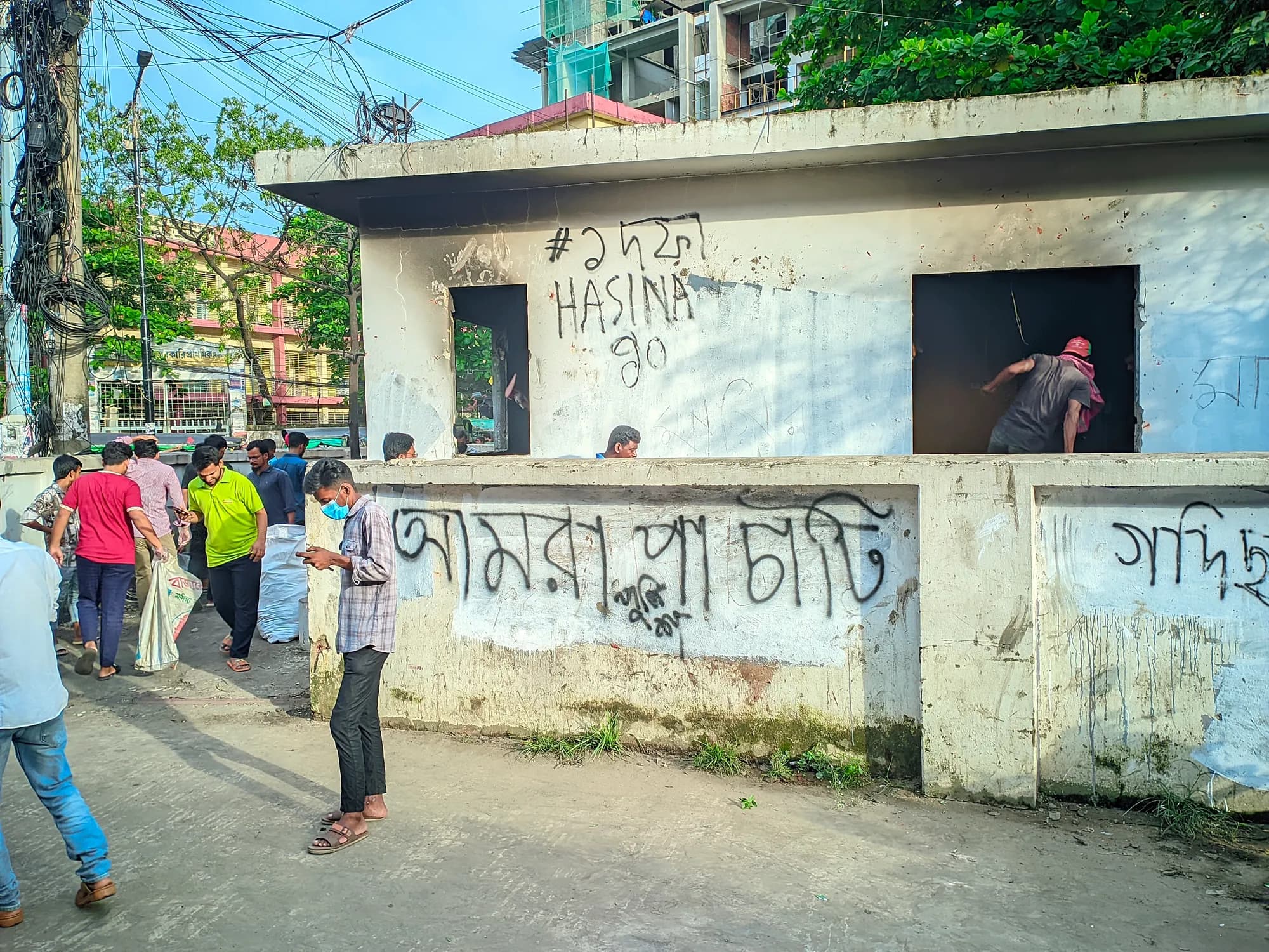 Young men work around a fire-blackened roadside police box covered in Bangla graffiti, one climbing through its window.