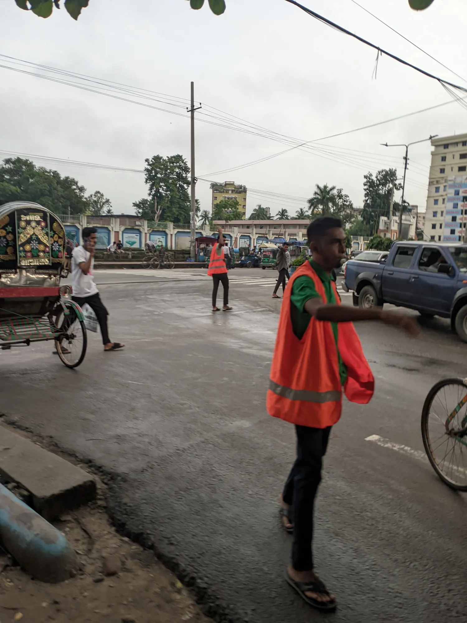 A young man in an orange vest signals with his arm on a wet road as a rickshaw and a cyclist pass.