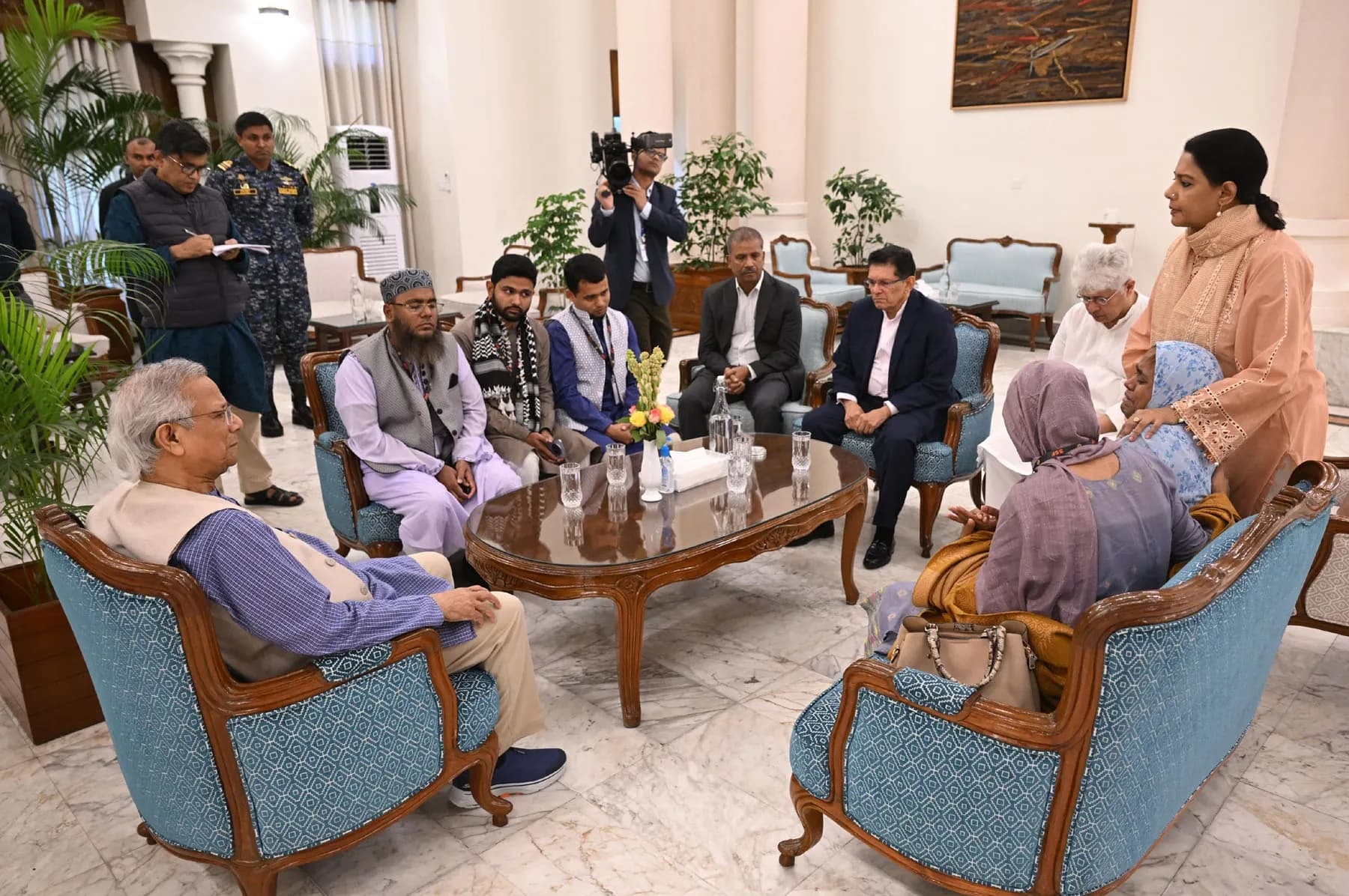 Several people sit in armchairs around a low table in a formal reception room while others stand behind them; a camera operator films.