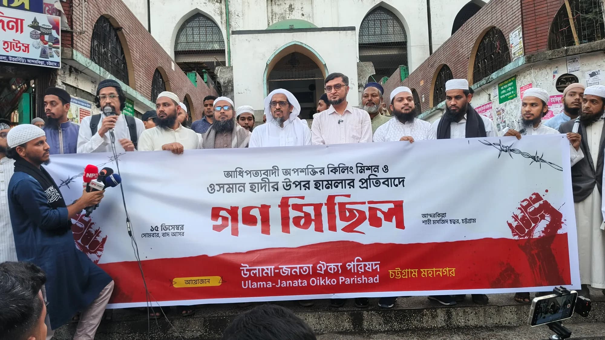 A line of men in white caps and panjabis stands behind a long red and white Bangla banner in front of an old mosque gateway.