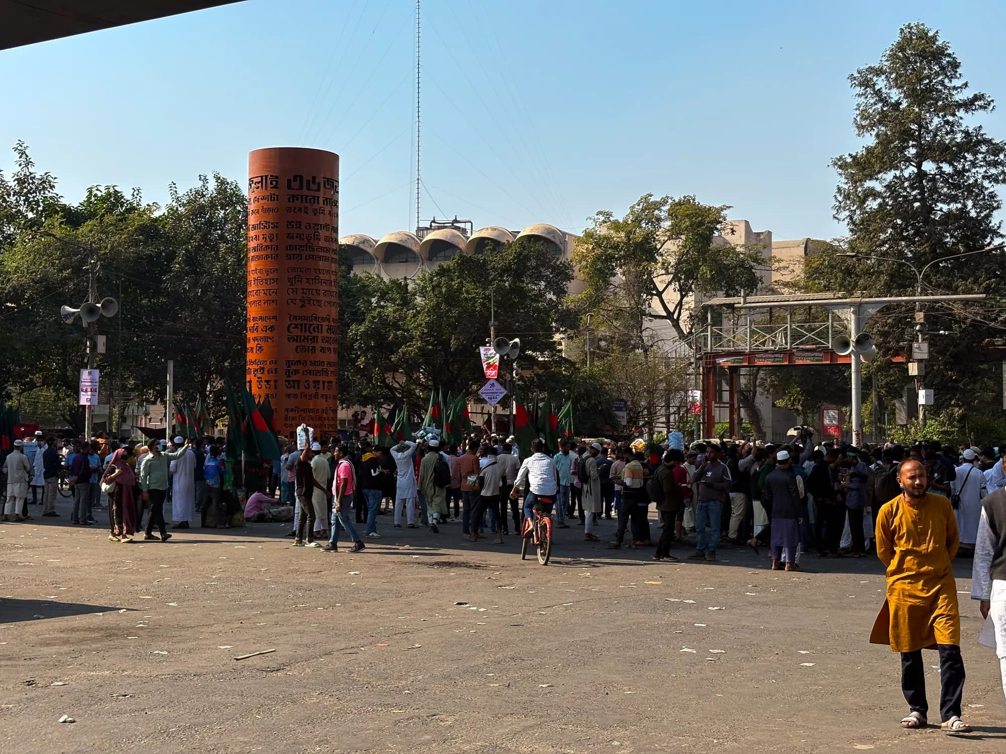 People stand in groups across a wide paved square beside a tall red brick column inscribed with Bangla text; national flags are held in the crowd.