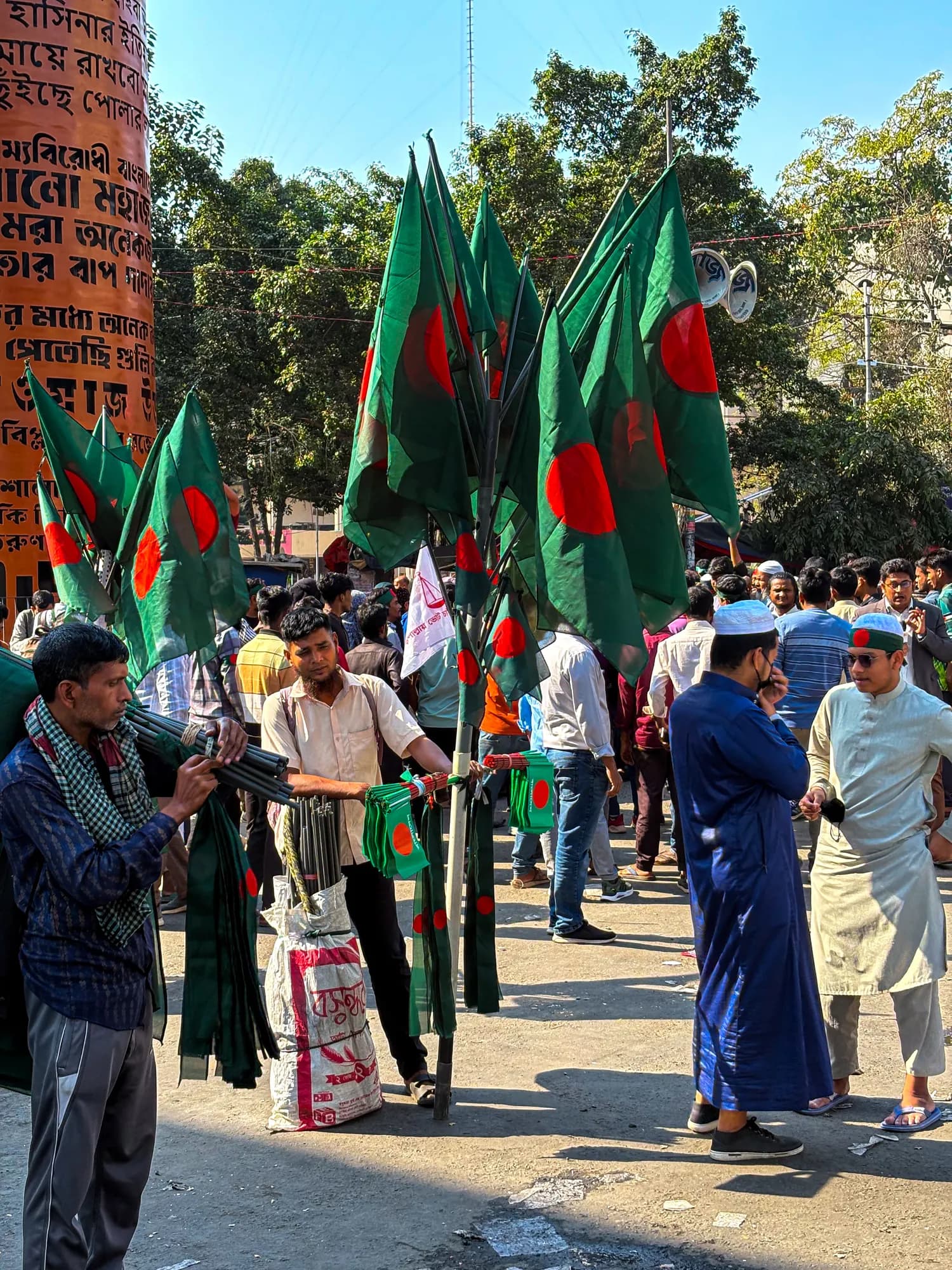 A man holds up a dozen green and red national flags on poles; people stand around him under trees on a sunny morning.