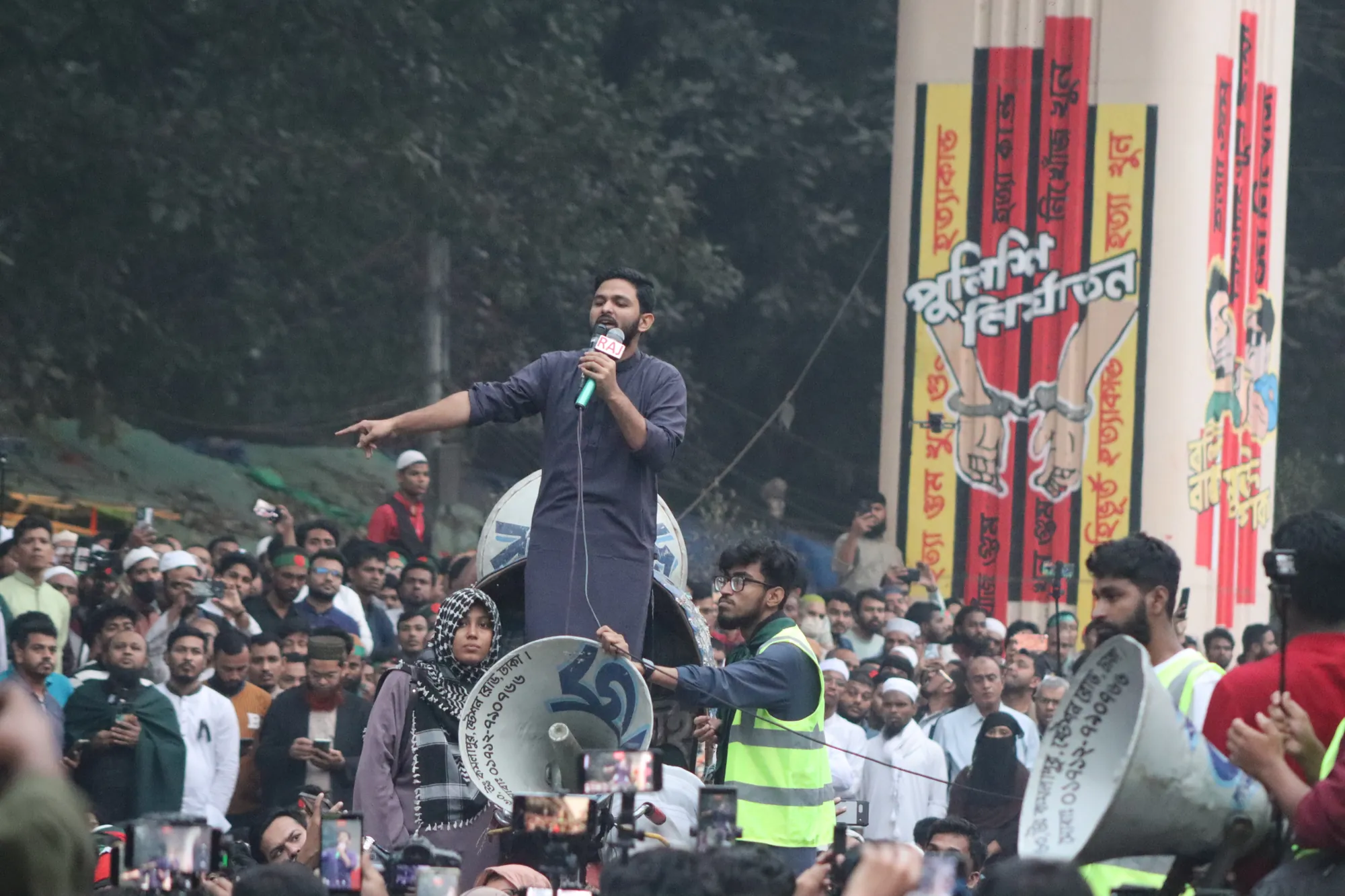 A young man in a dark kurta stands on a vehicle with loudspeakers, pointing as he speaks into a microphone above a dense crowd.