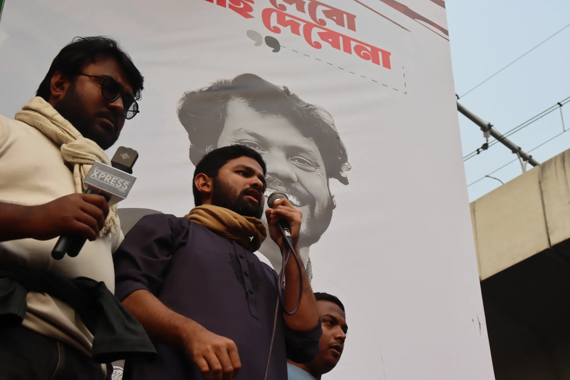 A bearded man in a dark kurta and scarf speaks into a microphone in front of a large banner with a printed face and red Bangla lettering.