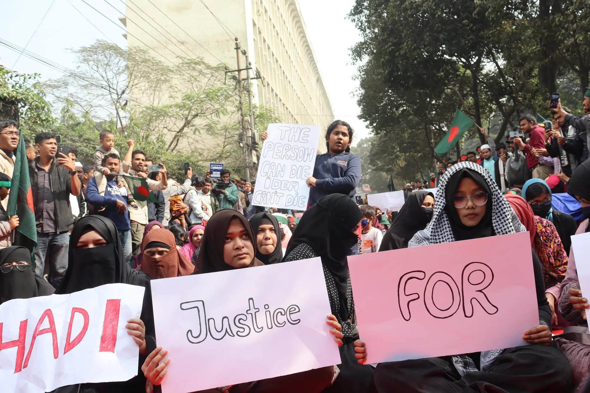 Women in headscarves seated on a road hold up pink and white hand-lettered placards reading “Hadi”, “Justice” and “For”.