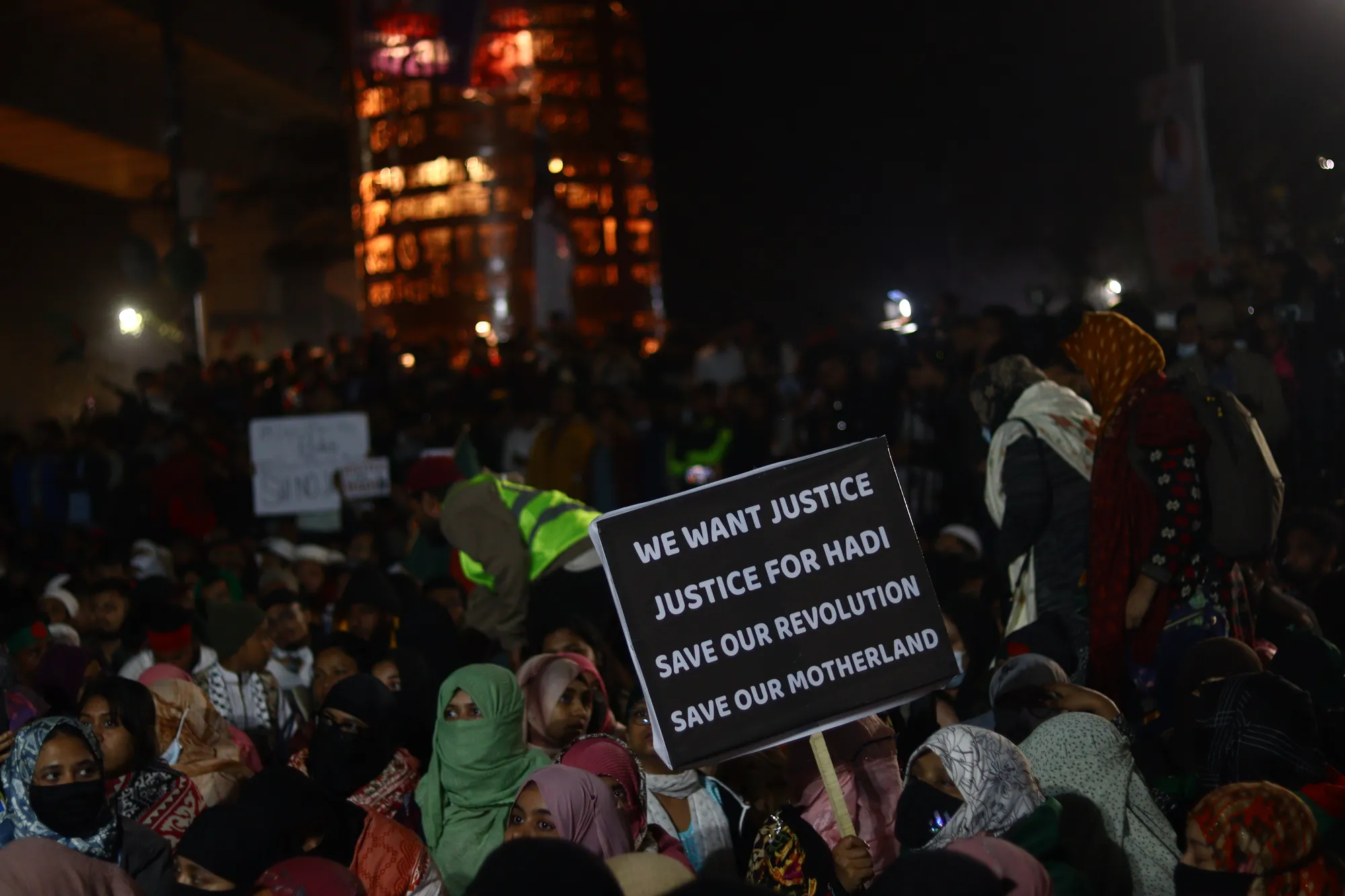 At night, a dark placard with white English lettering is held above a seated crowd, many in headscarves, lit by street lamps.