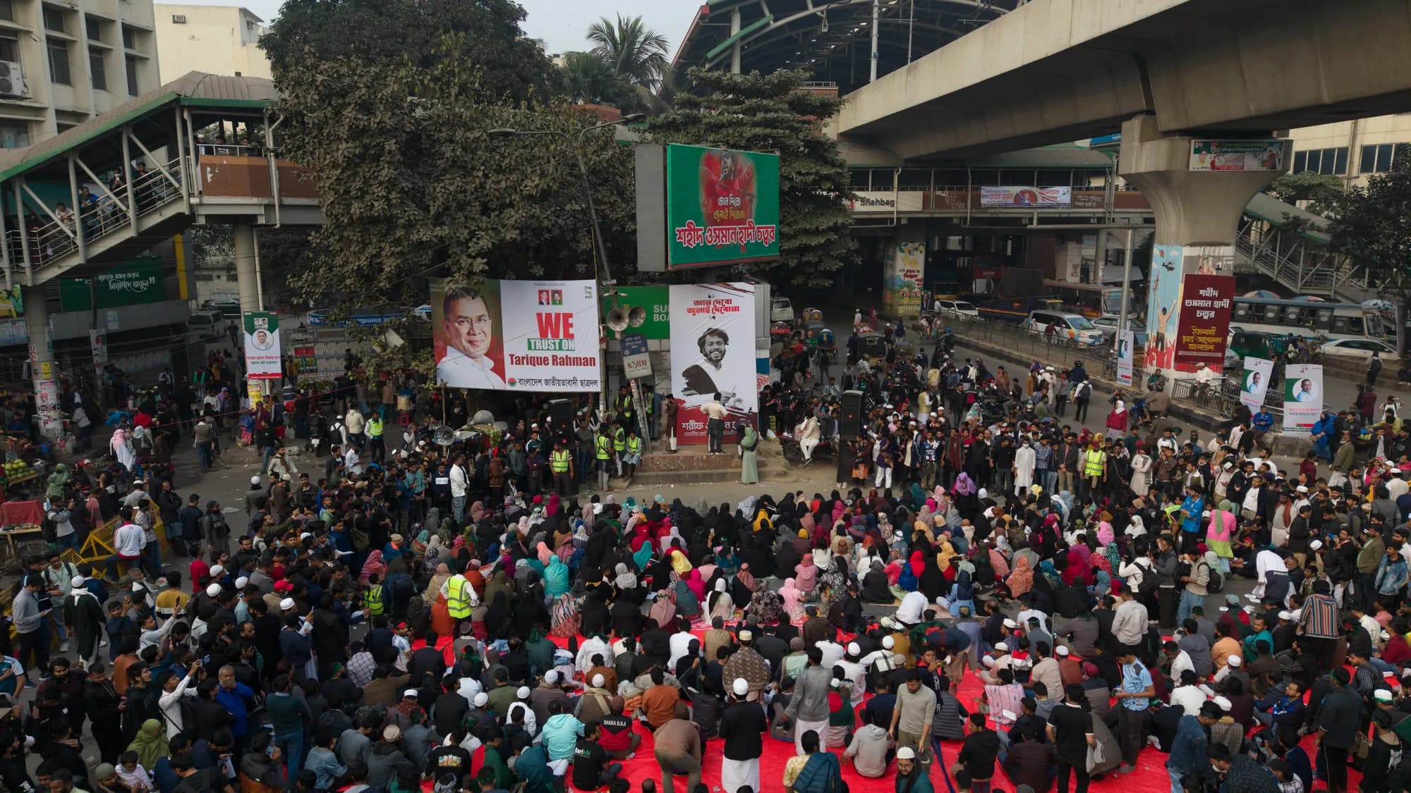 Seen from above, a large seated crowd fills a road junction beneath an elevated rail line, with banners and a stage at one side.