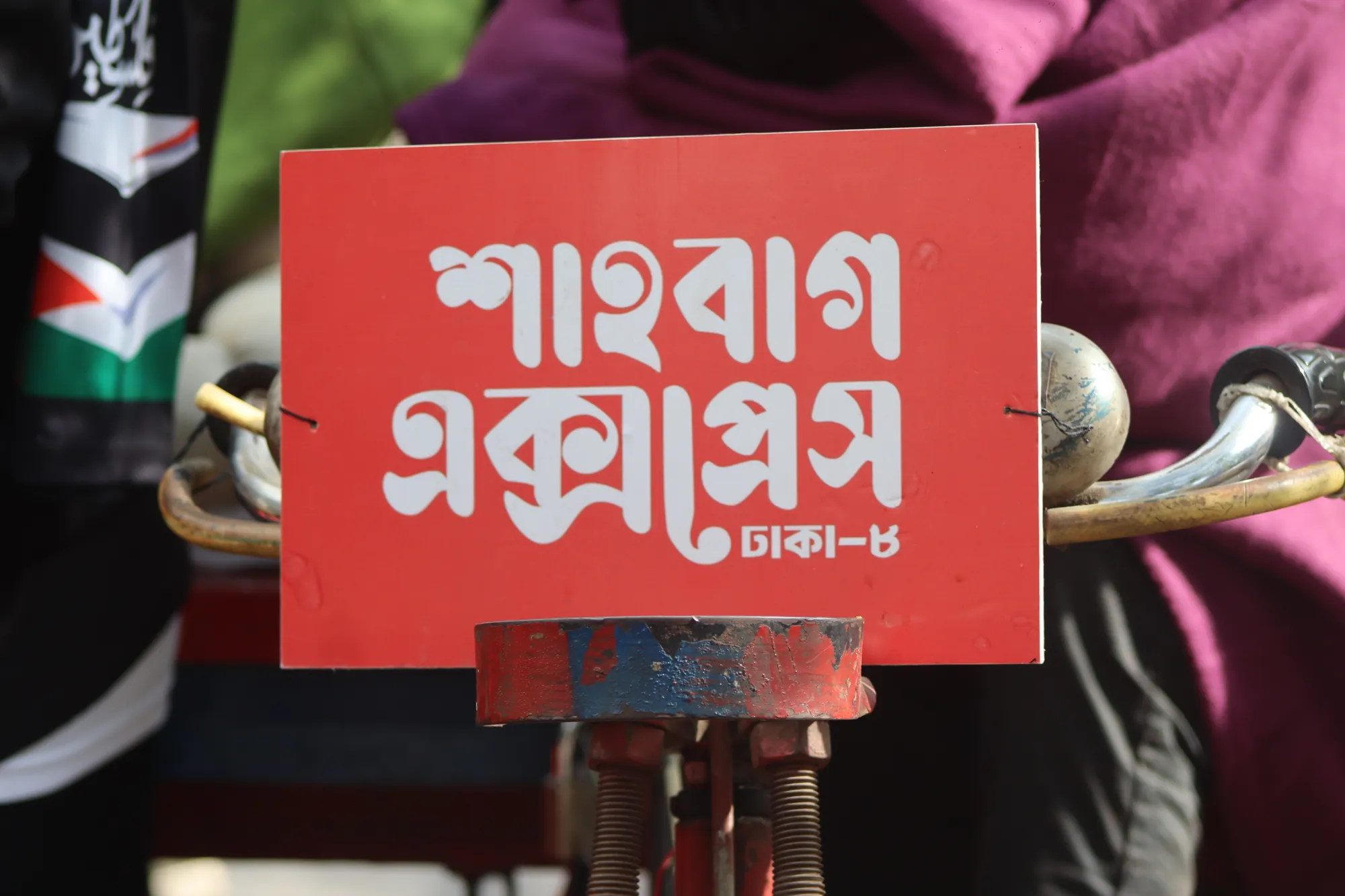 Close view of a red sign with white Bangla lettering mounted on a metal fitting, with fabric in the background.