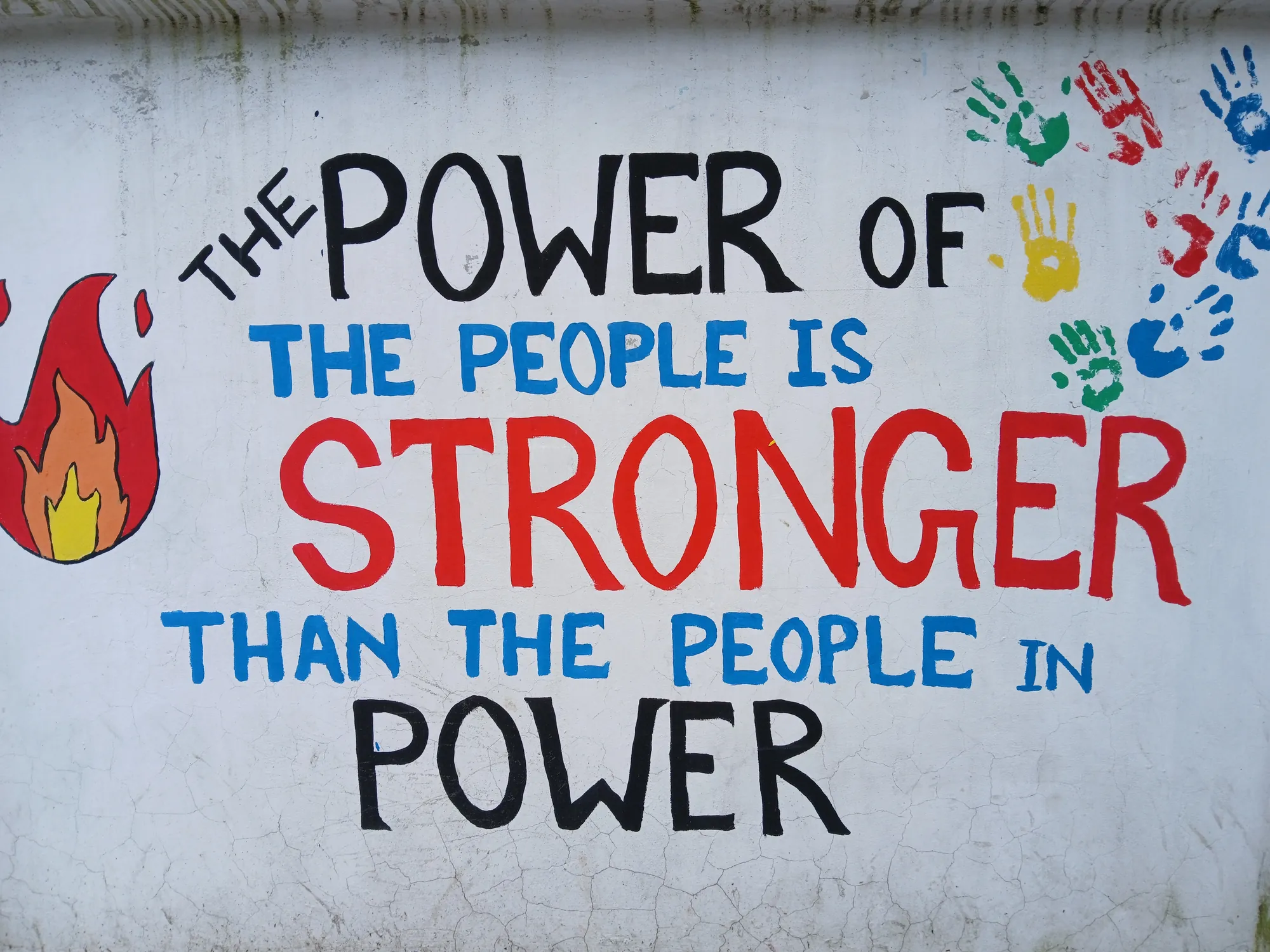 Hand-painted words “The power of the people is stronger than the people in power” in black, blue and red on a white wall, with coloured handprints.