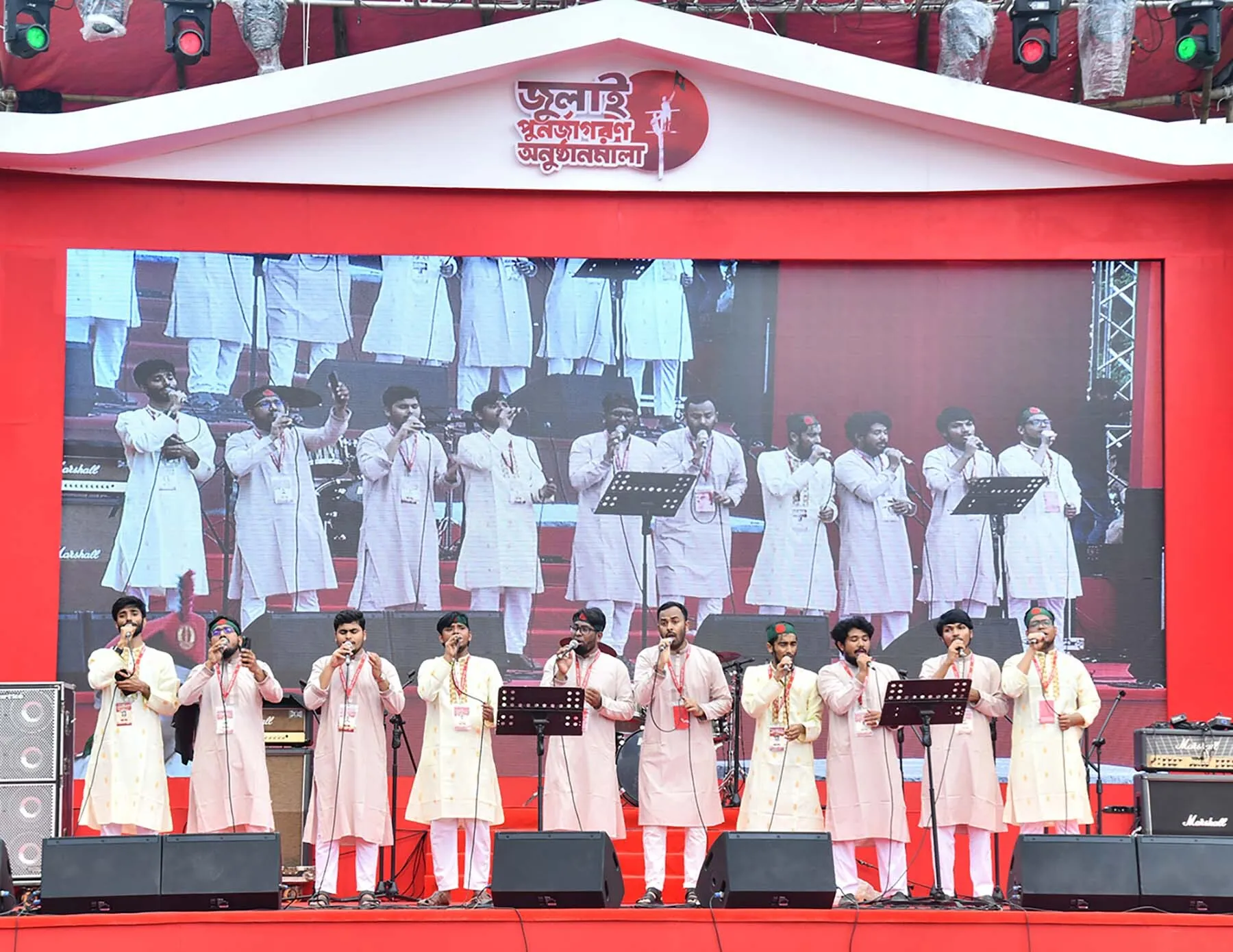 Two rows of singers in white panjabis perform on a red stage before music stands, with the same scene shown on a large screen behind them.