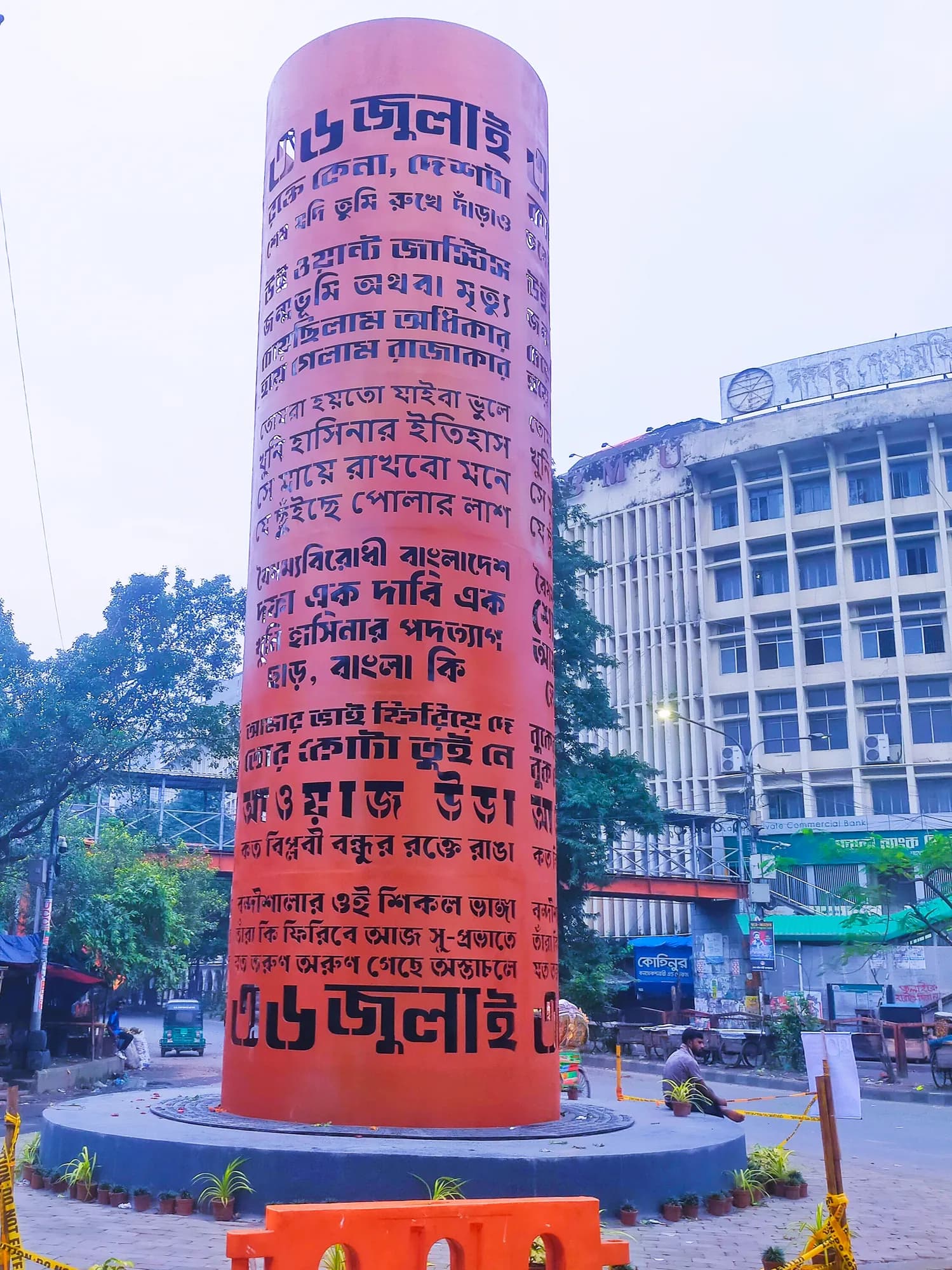 A tall orange-red cylindrical monument covered in black Bangla lettering stands on a plinth at a street corner before office buildings.