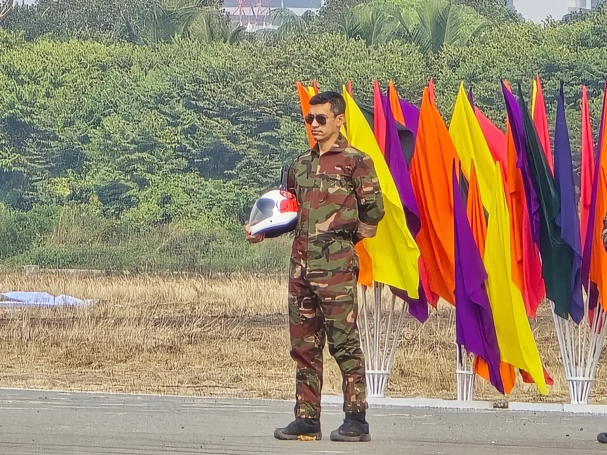 A man in a camouflage jumpsuit and sunglasses holds a white helmet at his side, standing before a row of tall coloured flags on an airfield.