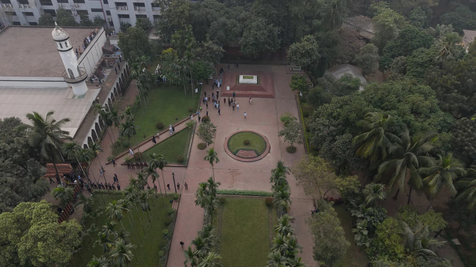 Aerial view of a mosque, a walled garden with paths and a round planted bed, and trees; small groups of people stand near a grave plot.
