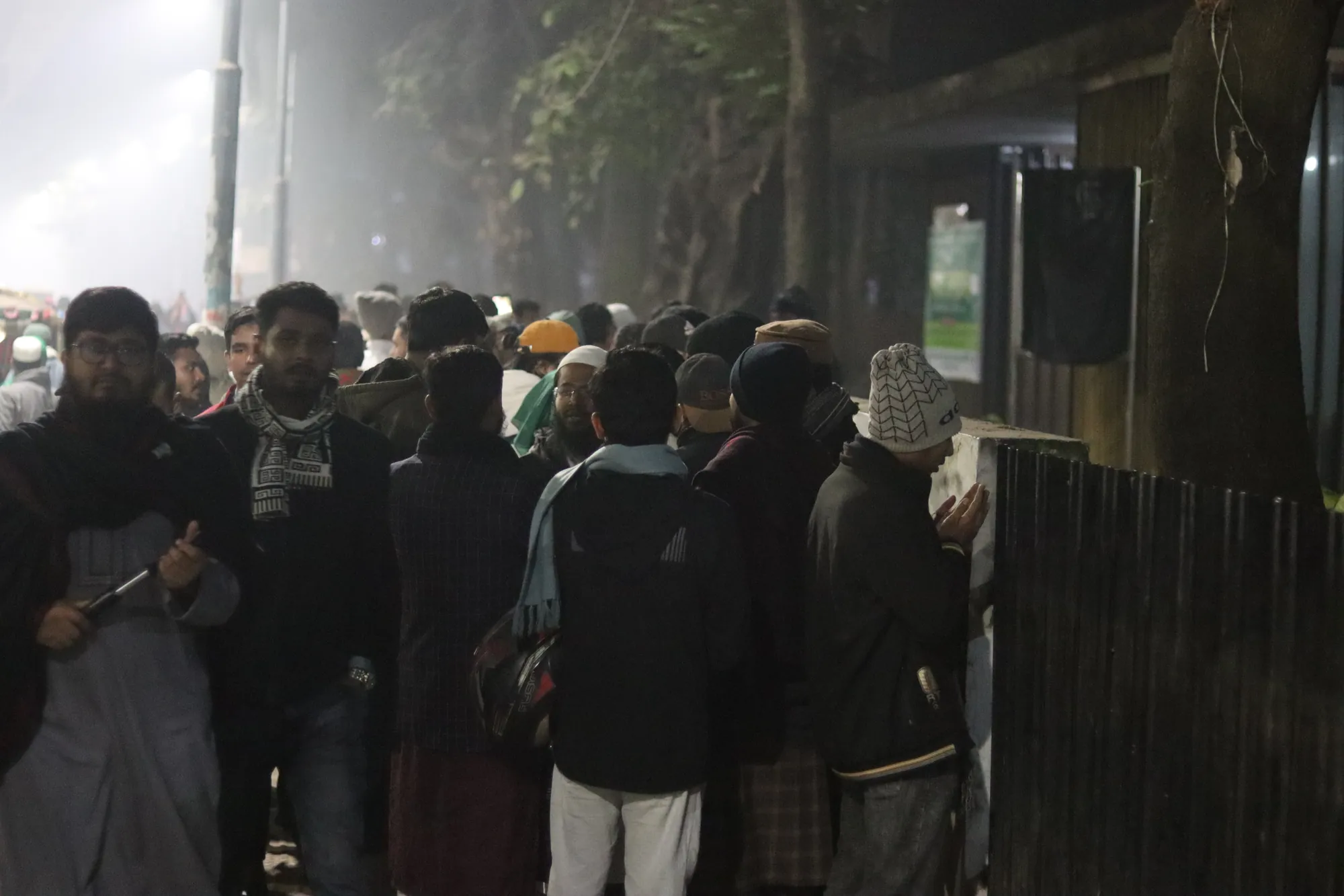 In dim light, men in winter clothes and caps stand by a dark fence; one raises his hands in prayer.