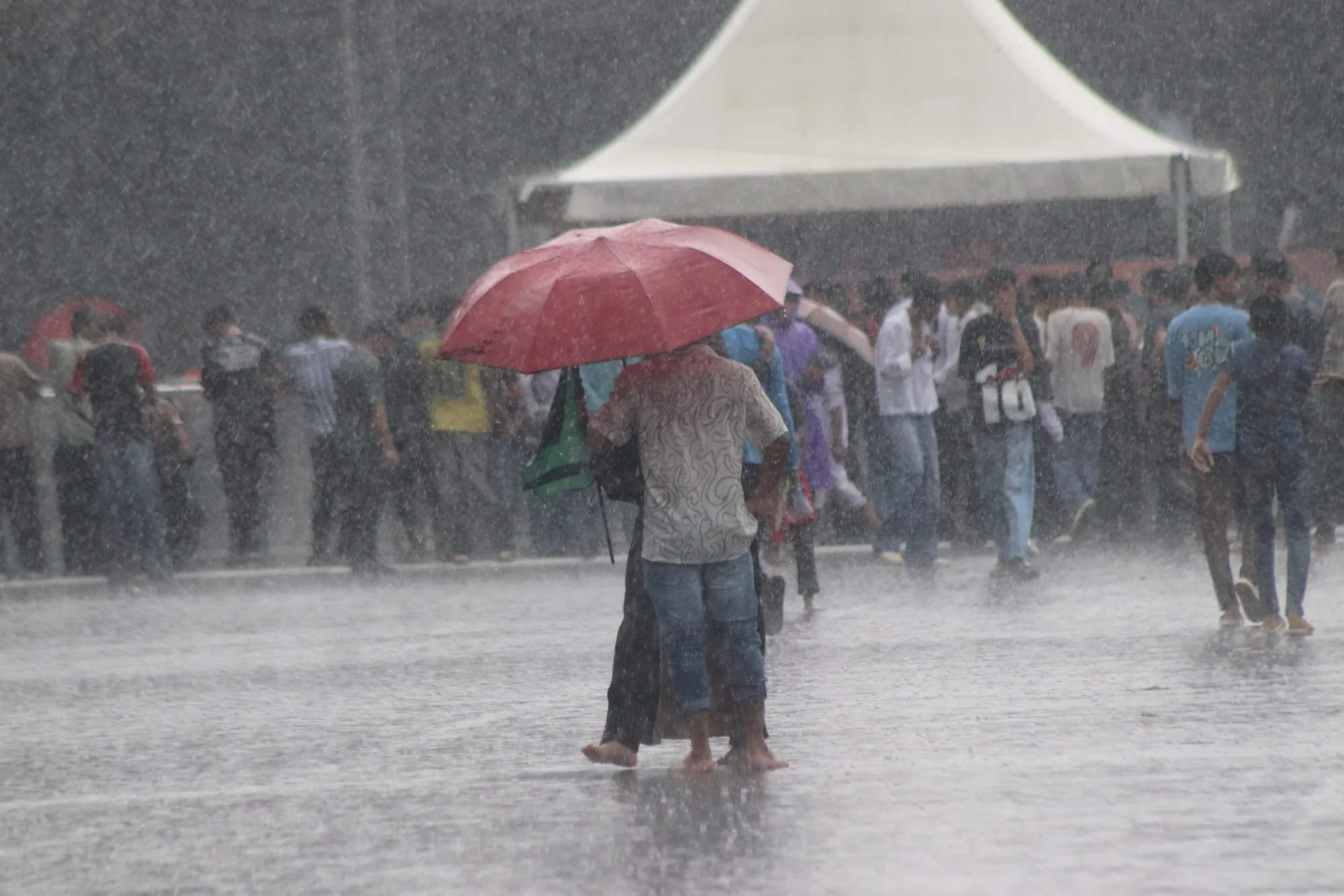 A man with a red umbrella stands barefoot on a flooded road in heavy rain; people shelter under a white tent behind him.