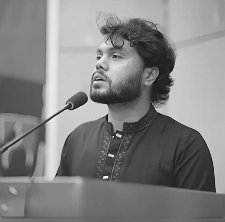Sharif Osman bin Hadi in a dark panjabi, speaking into a microphone at a lectern.
